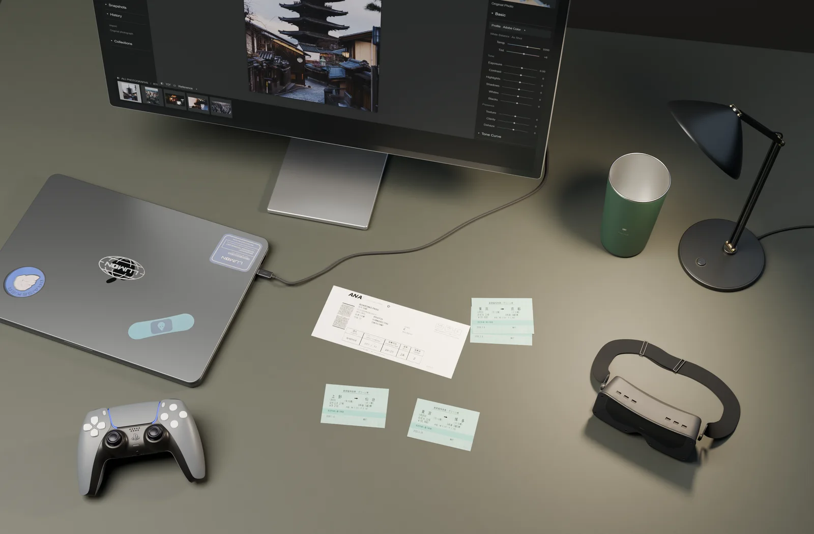 A warm lamp lights travel tickets on a finely textured grey-green desk, with a display, a 16-inch MacBook Pro bearing its Apple logo and four personal stickers, a controller, a dark-green cup and a Beyond headset.
